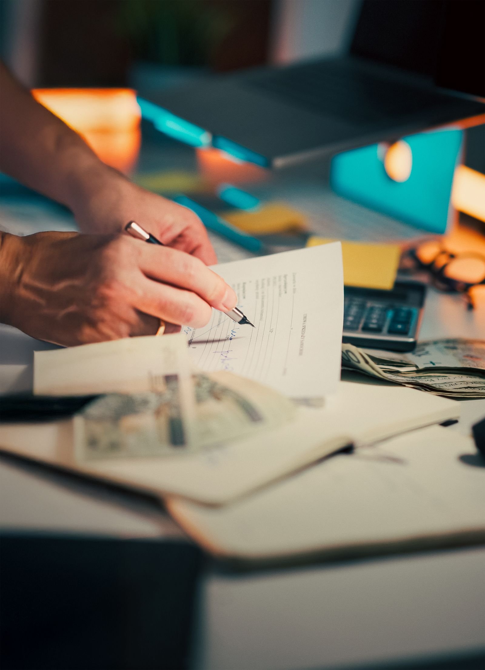 A hand signing financial documents on a desk with cash and a calculator, signifying finance management