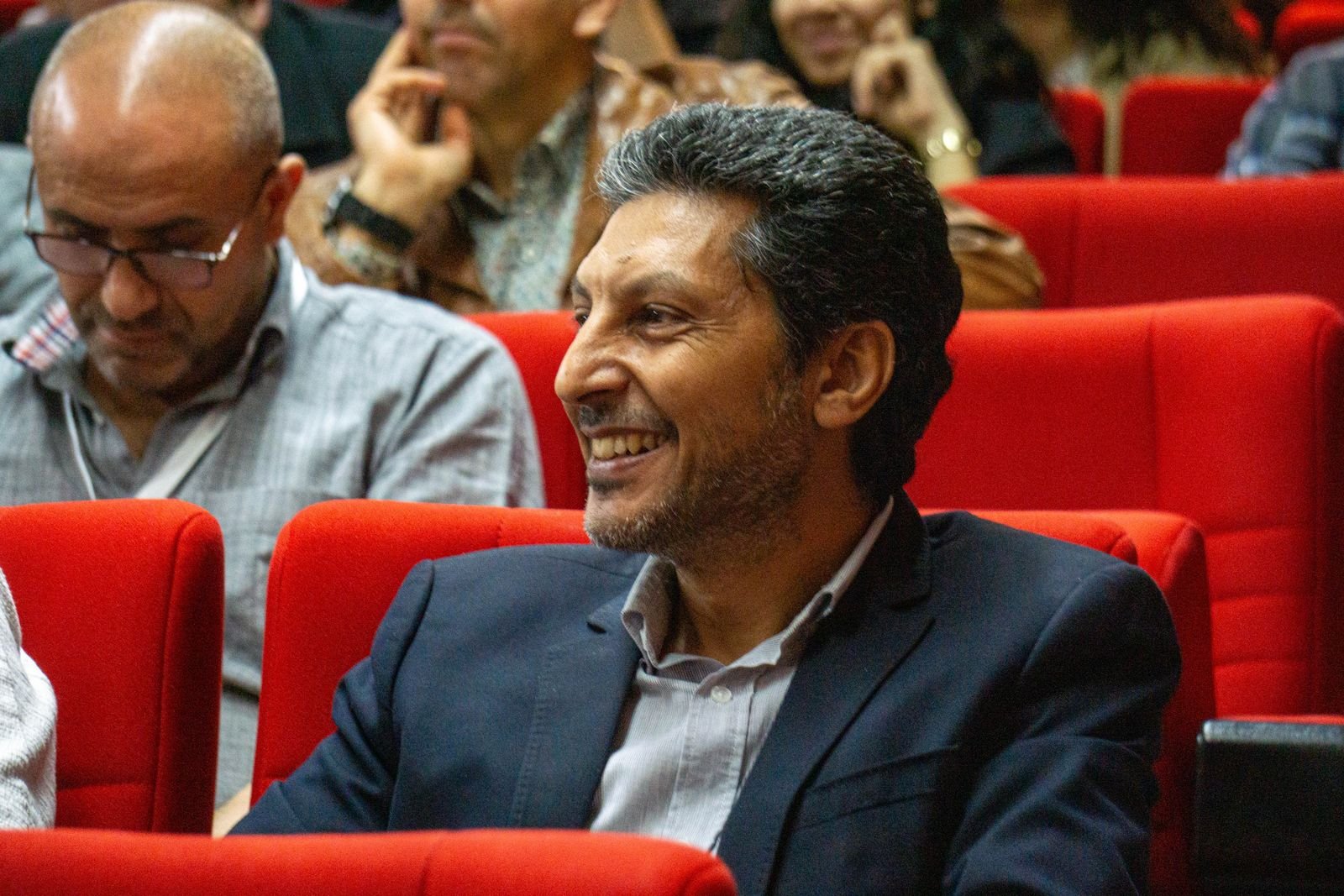 A man in a suit smiles while attending a conference, seated in a red auditorium