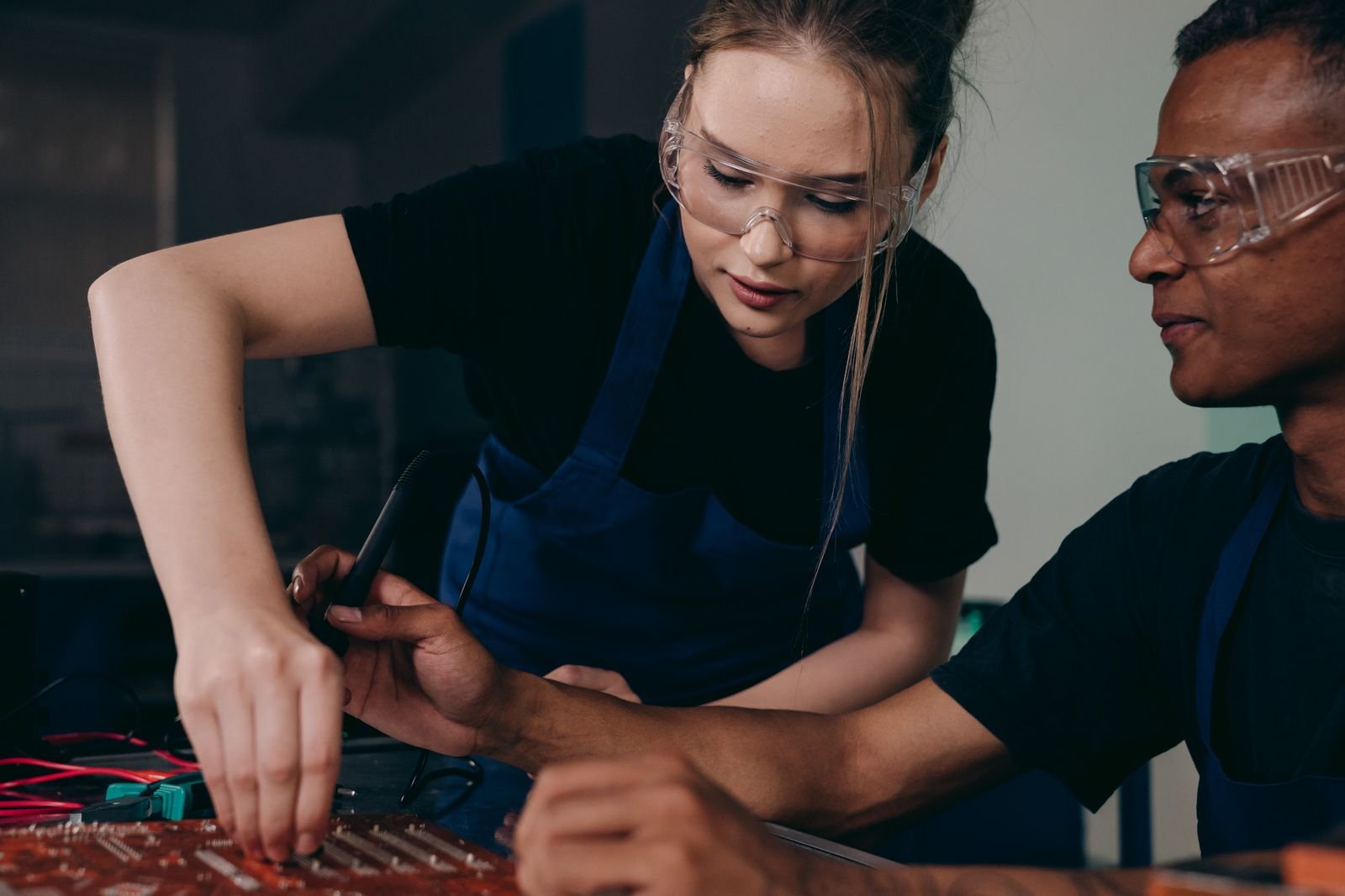 Two engineers wearing protective gear work together on a circuit board, focusing on technical precision